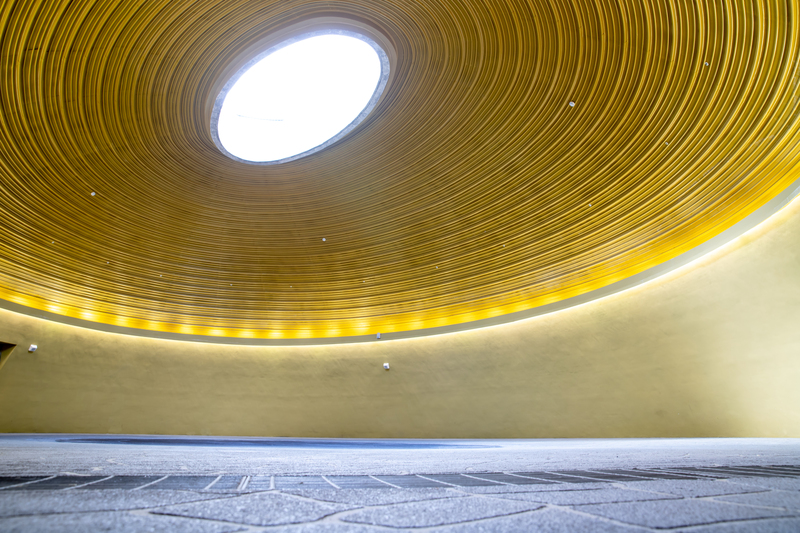 臺灣客家茶文化館的天光大廳室內空間，圓形挑高穹頂中央留有天窗，可透入自然光；金黃色的環形牆面與同心圓天花構成安定而開闊的場域，適合儀典與沉浸式表演。