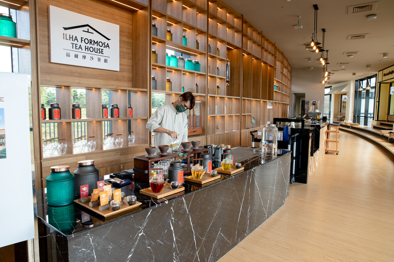 「福爾摩沙茶館內部茶吧台，工作人員現場示範熱茶沖泡與手沖流程，背景為整面茶罐展示牆，環境設計現代且具文化感，地點位於桃園龍潭的臺灣客家茶文化館